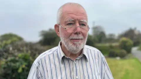 Hefin Wyn stands in a striped grey, white and blue shirt. Behind him is a garden and narrow lane. He has a beard and glasses.