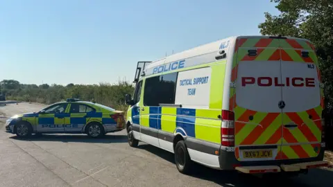 Leicester Media Online A police cordon in Derby Road, Loughborough on a sunny day. A police car and tactical support team van are parked outside the cordon. Two vehicles can be seen in the distance. 