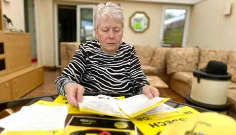 Martin Giles/BBC A woman wearing a black and white striped top sitting at a table laden with papers