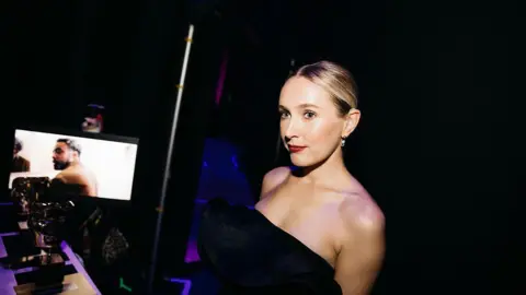 A glamorous woman in a black dress stands in the darkness behind a stage. 
