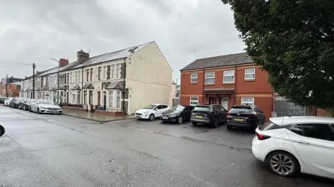 A long row of traditional stone and brick victorian terraced houses lines the left side of the street, featuring bay windows and small front gated areas. The other half of the street is modern built homes constructed from red brick and uPVC window frames.