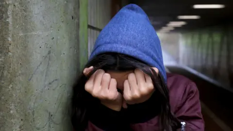 PA Media A head and shoulders image of a figure with a blue hood up and wearing a burgundy jacket. They hold their clenched fists to their face, obscuring their face. They are standing in concrete underpass, leaning against a wall. 
