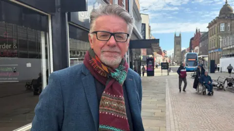 Martyn Cox poses for a picture on the high street with shops either side and church tower in the background. He wears dark glasses and a scarf with blue blazer.