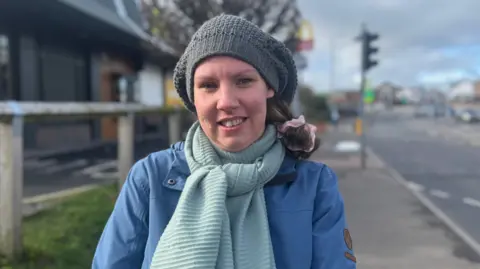 Ania Tabor. A woman wears a grey beanie hat and her brown hair is tied to the side. She is wearing a blue ribbed scarf and a darker blue raincoat. She is standing on a pavement on the side of the road in Glengormley.