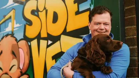 An image of veterinary co-owner James Buick. He is standing outside of a graffiti-style mural and smiling at the camera. He is holding a brown cocker spaniel dog. James has short brown hair and is wearing a blue jacket.