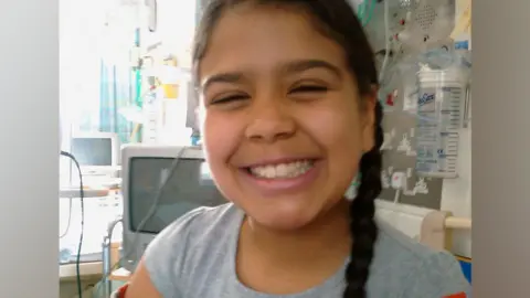 Family A close up of Jasmine, who is wearing a grey top and is smiling broadly at the camera. Behind her is a television on a stand and various hospital equipment