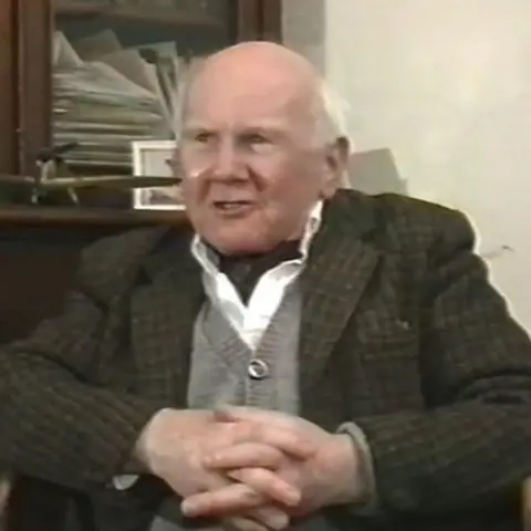 Ron Fairfax A man with a balding head and tufts of white hair sits in front of a dark-wood, glass-fronted cupboard with his hands crossed in front of his stomach. He is wearing a dark tweed jacket over a grey cardigan, white shirt and dark cravat.