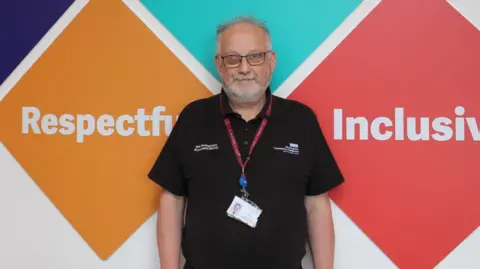 Birmingham Community Healthcare NHS Trust Keith has grey hair and black glasses, and is wearing a black t-shirt with the hospital trust logo on the breast in white. He has a red lanyard around his neck which has a photocard on the end. A wall behind his back has colourful diamond shapes, with white writing that reads " Respectful" and "Inclusive"