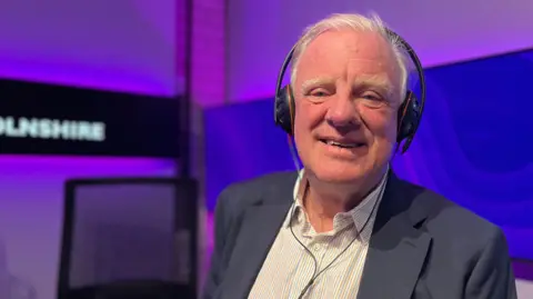 Sir Edward Leigh wearing a navy blazer and a white striped shirt with a pair of headphones. He has white hair and is smiling at the screen. He is sitting in a radio newsroom and the background is purple. 