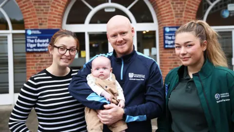 University of Worcester Two women standing either side of a man who is holding a baby. On the left is the baby's mother, who has ginger hair tied back and is wearing glasses and a black and white striped top. The man, the baby's father, is bald with ginger facial hair and is wearing a navy top that says "university of Worcester" on it. on the right is a student paramedic who has light brown hair tied in a ponytail. she is wearing a teal t-shirt and jacket, with the university of Worcester logo on it