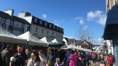 Cumberland Council Keswick Market in its modern iteration. There are dozens of people milling about market stalls which have large gazebos saying "Cumberland Council" sheltering them. A hotel and a pub can be seen behind the stalls.