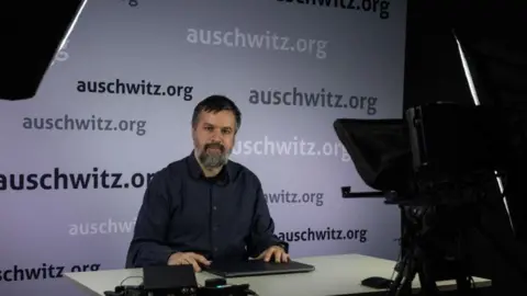 Cilvēks ar bārdu ar melniem un pelēcīgiem matiem sēž uz galda ar slēgtu klēpjdatoru. Fonā ir siena, kurā vairākas reizes ir uzrakstīts vārds auschwitz.org. Viņš ieskatās kamerā