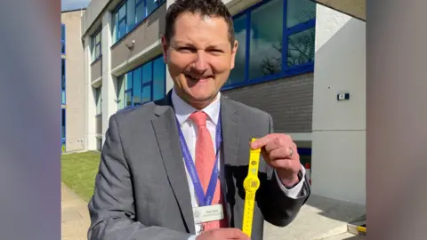 Dave Savill wearing a grey suit, white shirt and a pink tie. He is holding a yellow watch-like wristband with both his hands.