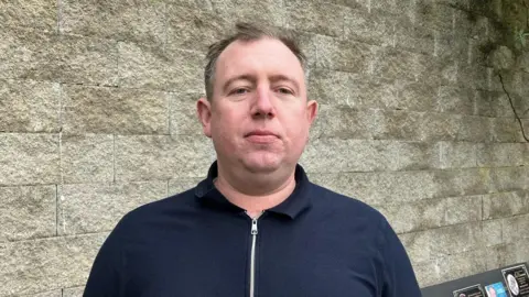 The picture shows a man with short thinning brown/grey hair in a navy zip-up top