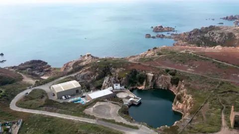 BBC An aerial view of the desalination plant off the coast. A road can be seen winding down towards a large building with cars parked outside it and a body of water. Rocky surroundings face the ocean.
