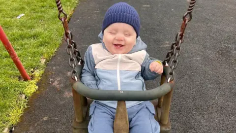 Stepan Kozariichuk Bohdan is smiling brightly while sitting in a playground swing. His eyes are closed and his hand is raised. He is wearing a blue and white coat and a blue hat.