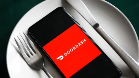 A mobile phone showing the DoorDash logo sits between a fork and a knife on top of a dinner plate