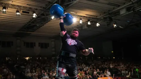 Crown Copyright 2026 Rhys Calland lifting a heavy weight in his right hand in front of a crowd of people at a competition.