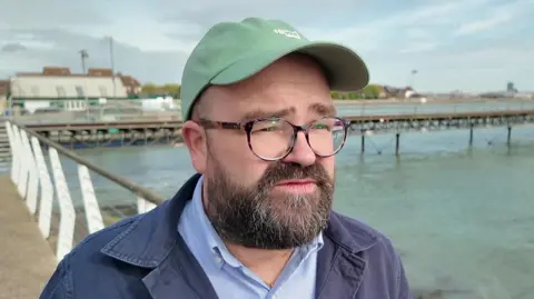 A man with a dark beard is pictured in front of a pier and a body of water. He is wearing a green baseball cap and glasses. He is also wearing a dark blue jacked over a light blue collared shirt. 