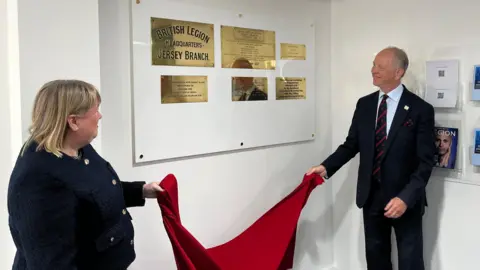 BBC The image shows two people holding some red fabric unveiling a series of commemorative brass plaques. They are in a room with white painted walls.