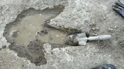 A single track dinosaur footprint. The large print is full of greyish water with a trowel in one of the three toes. The print is in a grey stone. There is a black glove in one corner of the photo.