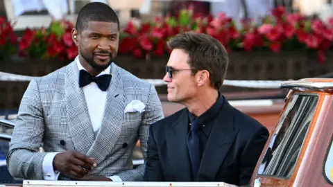 Getty Images US singer-songwriter Usher Raymond IV (L) with American football star Tom Brady - Usher wears a grey check suit and black bow tie, Brady wears a black suit, dark navy tie and dark glasses, and both are aboard a boat.