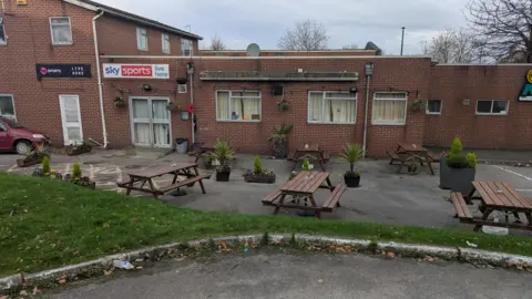 David Spereall/BBC Some empty picnic tables sit outside a club made of bricks. 
