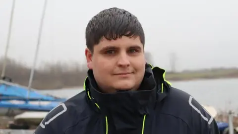 A young man stood looking at the camera, with sailing boats and a water body slightly visible in the background.