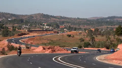 A wide shot showing the finished road stretching into the distance. Cars can be seen travelling on the pristine tarmac.