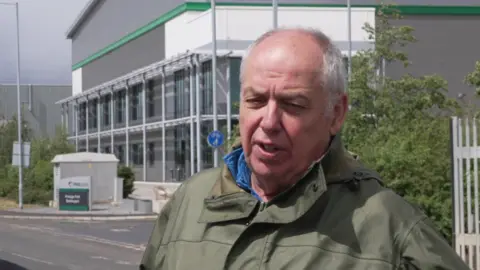 Nick Matty with grey hair, wearing a green hooded jacket, speaks during an interview outdoors, with a large grey and white commercial building and a 'Prologis Park Beddington' sign behind him