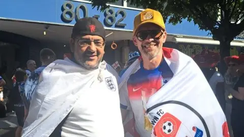 Two men wearing flags wrapped round their shoulders smile at the camera