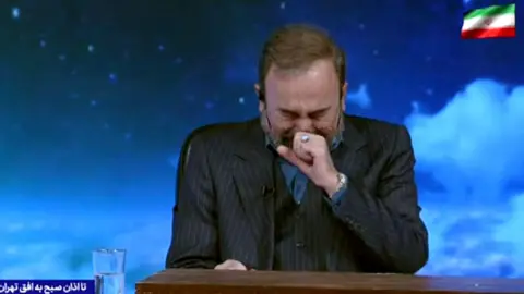 Image shows a presenter on TV wearing a dark suit, with one hand to his face, eyes closed, and crying. The background behind him shows a blue sky with clouds
