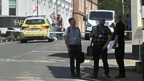 Three men standing in an empty street in shadow with a police car and a row of white buildings in the background as well as a police car
