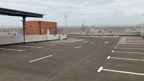 A view of empty spaces on  the top floor of the car park, surrounded by a high metal mesh fence.