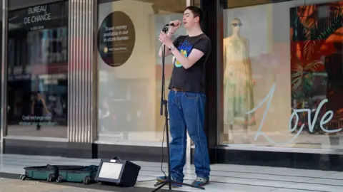 BBC Person singing into a microphone on a stand while busking outside a row of city centre shopfronts. An amplifier and open cases sit on the pavement in front of the performer, with store windows and signage in the background.