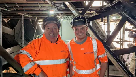 Paul and Charlie, wearing orange high vis clothing and dark hard hats, stand smiling at the camera with the steel beams of the bridge and scaffolding stretching out from all angles around them. 