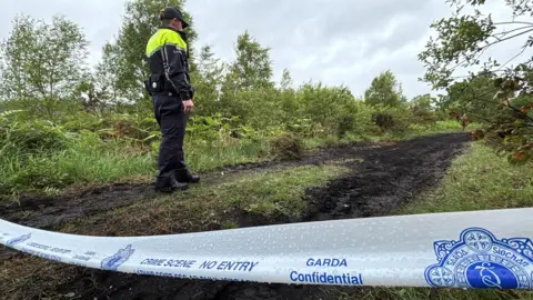 Niall Carson/PA Wire A garda officer standing near a white cordon during a search of open ground outside Tullamore, County Offaly, on Monday 26 May 2025. The officer is wearing a cap and a dark uniform with a yellow high-vis section around his shoulders and chest. He is looking towards a dark, boggy lane, surrounded by green vegetation.