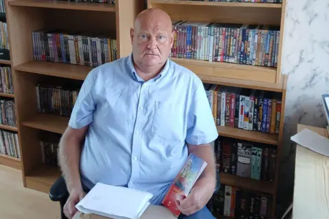 Mark Corfield Mark Corfield is sitting in front of a bookcase full of DVDs with a pile of parcels on his lap.