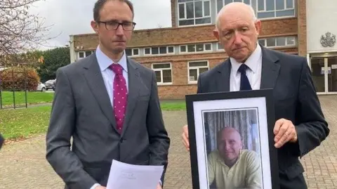 Marc Gaier/BBC Amos Waldman, who has glasses and brown hair and wears a grey suit with a pink tie, and Dr Steven Waldman, look at the camera with sorrowful expressions while holding a framed photo of a smiling Dan Kay