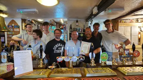 Staff at The Nag's Head are pictured behind the bar. There are eight male staff members pictured. In front of them are various beer taps and one man, wearing a black top is holding a white certificate. 