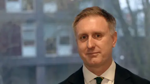 John Fairhall/BBC Ed Garrett, a man with short light brown hair wearing a shirt and tie. He is looking into the camera. 