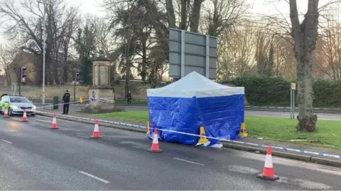 Tom Percival/BBC Forensic scene guard (blue tent) on the edge of a pavement by a busy roads, with road cones protecting it