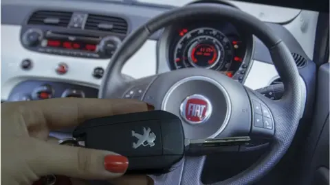 Anadolu Agency / Getty Images A woman holds a Peugeot car key in front of a Fiat steering wheel