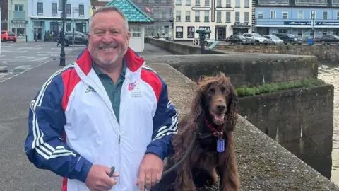 Holidays for Heroes Jersey Mark smiles at the camera as Jerry sits on a wall next to him and Gorey is in the background