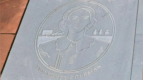 An image of Ann Raney Coleman etched on to a flagstone. The design shows a head and shoulders image of a woman with short hair. Behind her are three men on horses and a horse-drawn cart.