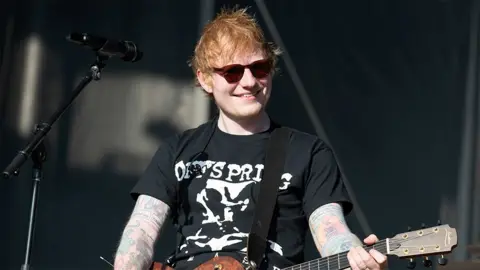 Getty Images Ed Sheeran, smiling and wearing a black tshirt with white writing, brown sunglasses, performing on stage with a guitar in hand and a microphone on a stand next to him.