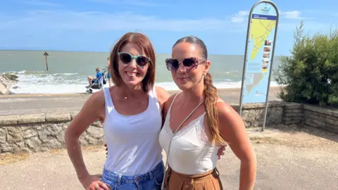 Jaimielee Rendall/BBC Two women in white T-shirts and sunglasses stand on the seafront with an arm around each other. Both are smiling. The sea is in the background