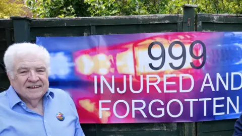Tom Curry Tom Curry in a blue shirt standing beside a blue and red banner saying "999 INJURED AND FORGOTTEN".