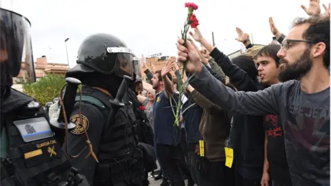 Getty Images Crowds raise their arms up and a man holds red and pink flowers as police move in on members of the public gathered outside to prevent them from voting in the referendum at a polling station where the Catalonia President Carles Puigdemont will vote later today on October 1, 2017 in Sant Julia de Ramis