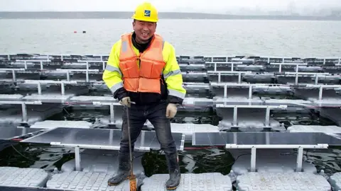 BBC Wu Wenhui in yellow hard hat and orange life jacket standing on floating solar panels